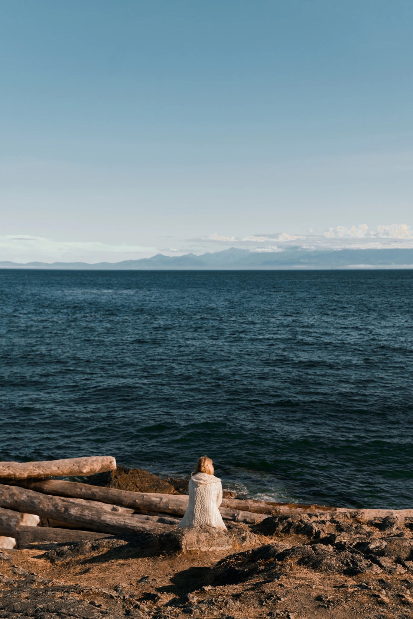 Woman in a knitted sweater sitting on a rocky shoreline looking out over the ocean, representing Revalio Health’s origin story and commitment to everyday well being.