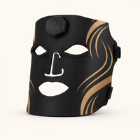 Revalio black silicone LED facial therapy mask with gold wavy accents and white cutouts for the eyes, nose, and mouth, shown on a light cream background.