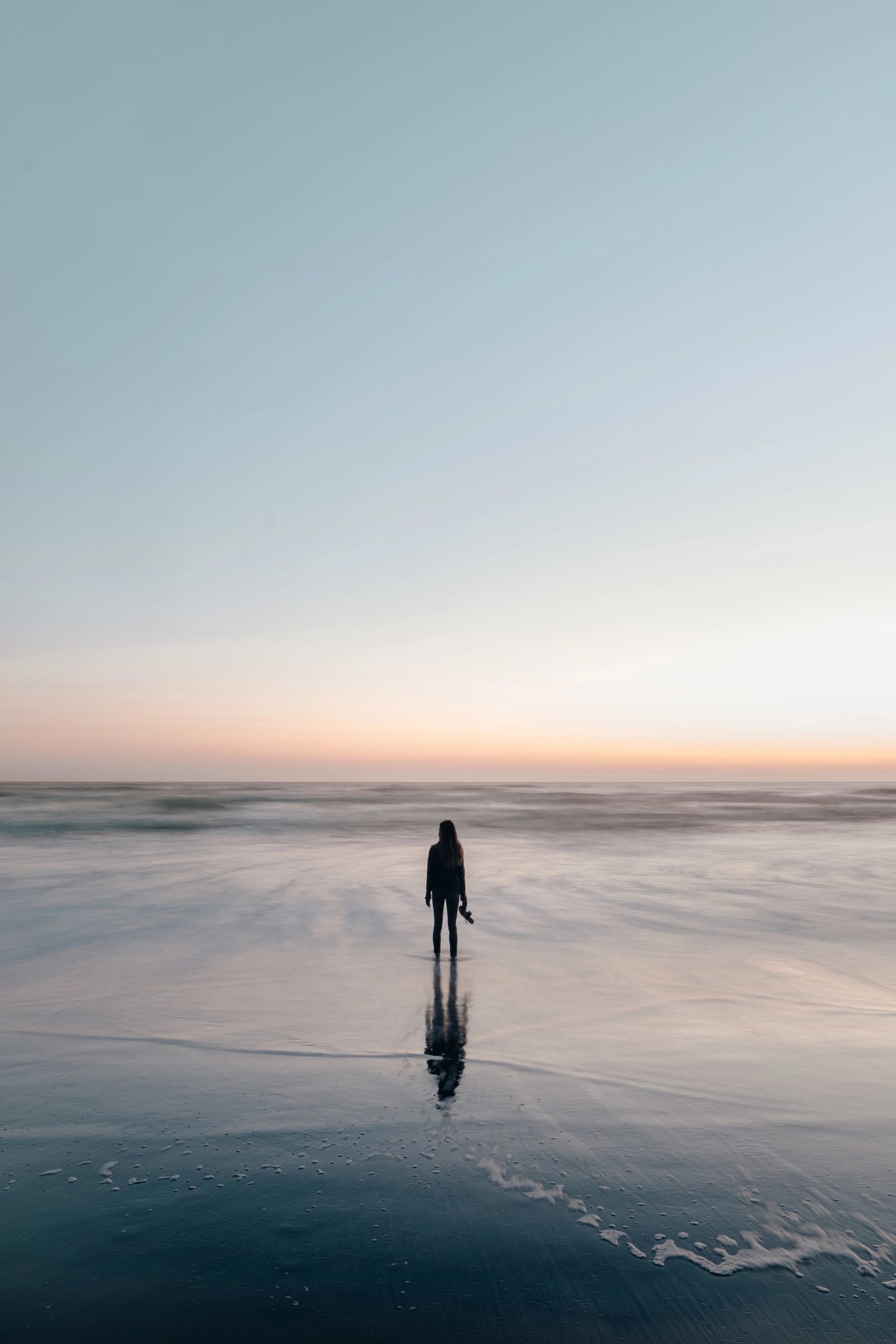 A woman standing at the shoreline during a calm sunset, looking out at the ocean; a peaceful hero image representing wellness and transformation.