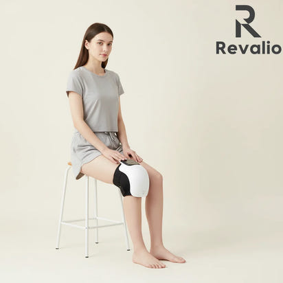 Woman using the Revalio Smart Knee Massager With Heat while sitting on a stool with Revalio logo in the corner.
