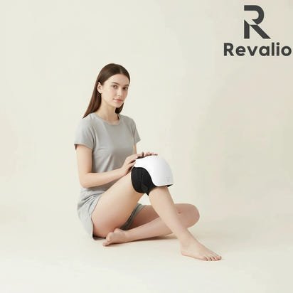 Woman using the Revalio Smart Knee Massager with heat and red light technology while sitting the floor with Revalio logo in the corner.